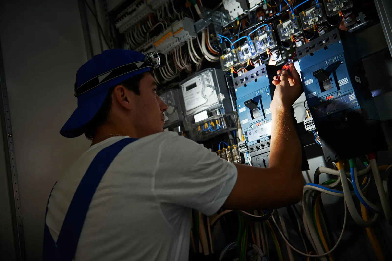 Electrician in blue cap works on wires inside a dimly lit electrical panel, focused expression, vibrant colored cables visible.