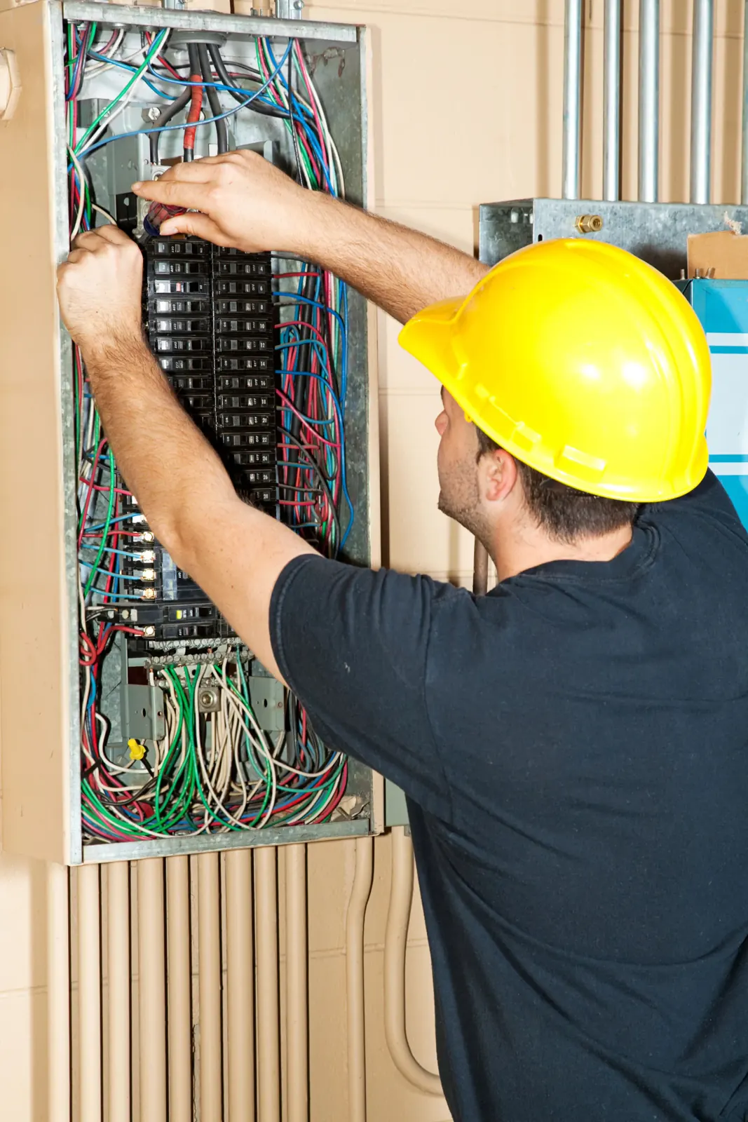 Electrician in a yellow hard hat working on a breaker panel with colorful wires, set indoors against a beige wall.